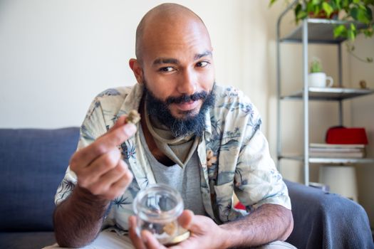 Smiling bald man with beard holding cannabis at home with a relaxed and cheerful expression.