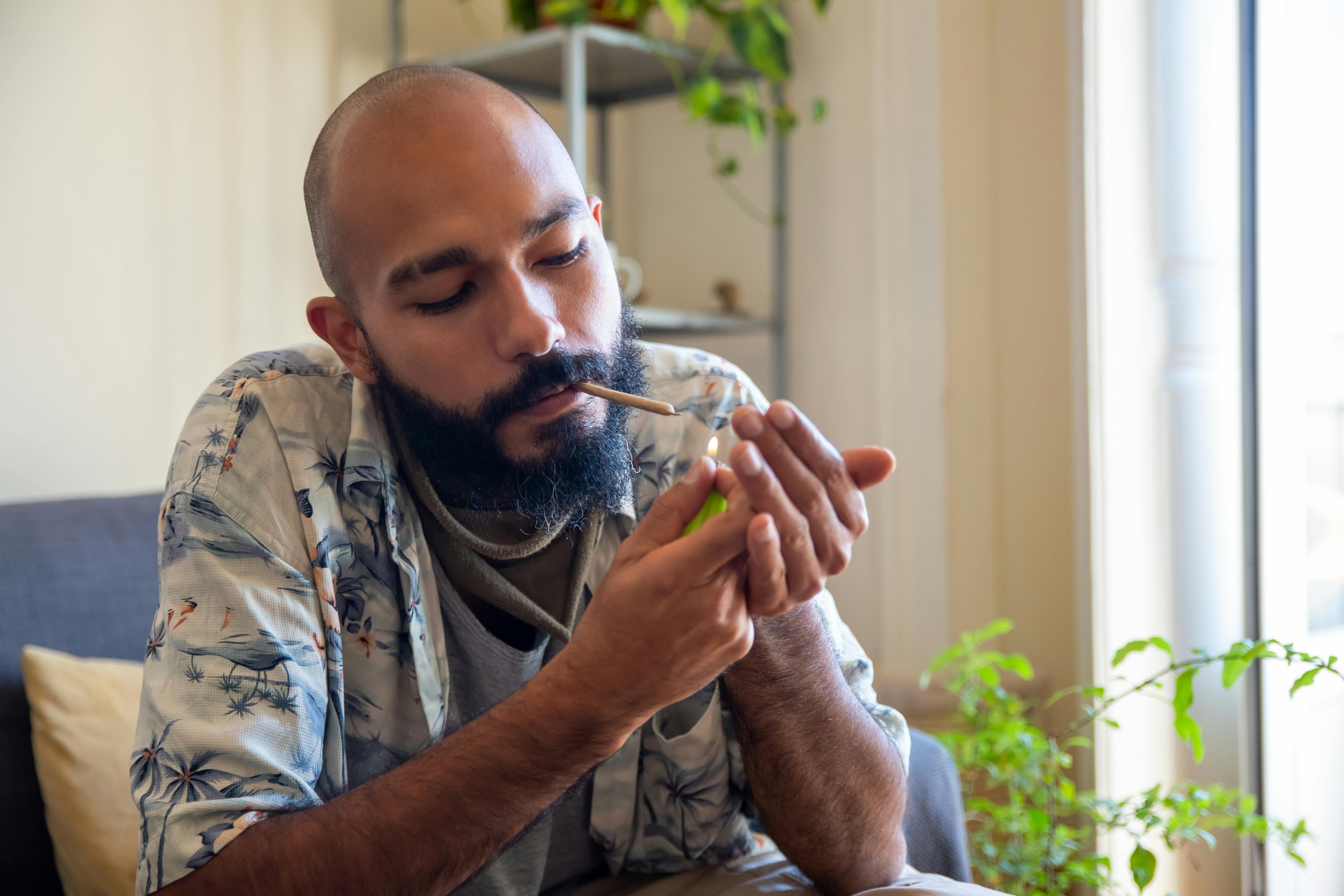 Bald man with beard lighting a cannabis joint indoors, captured in a cozy setting.