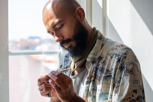 Bald bearded man in a printed shirt holding a joint near a sunlit window indoors.