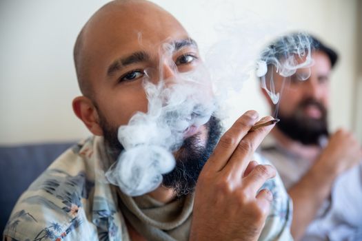 Close-up of a bearded man smoking cannabis indoors with a friend.
