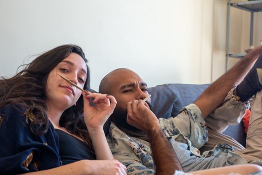 A couple relaxes on a sofa, playfully pretending to smoke, embodying a casual, fun atmosphere.