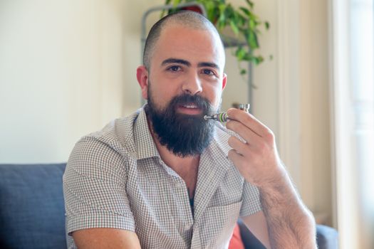 A bearded man indoors enjoying a smoke from a stylish pipe, exuding calm and relaxation.