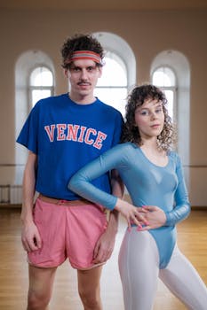 Two people in vibrant 80s fitness attire posing indoors.