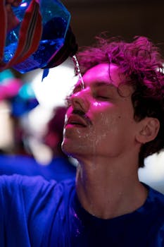 Sweaty man pouring water on his face under vibrant neon lighting.