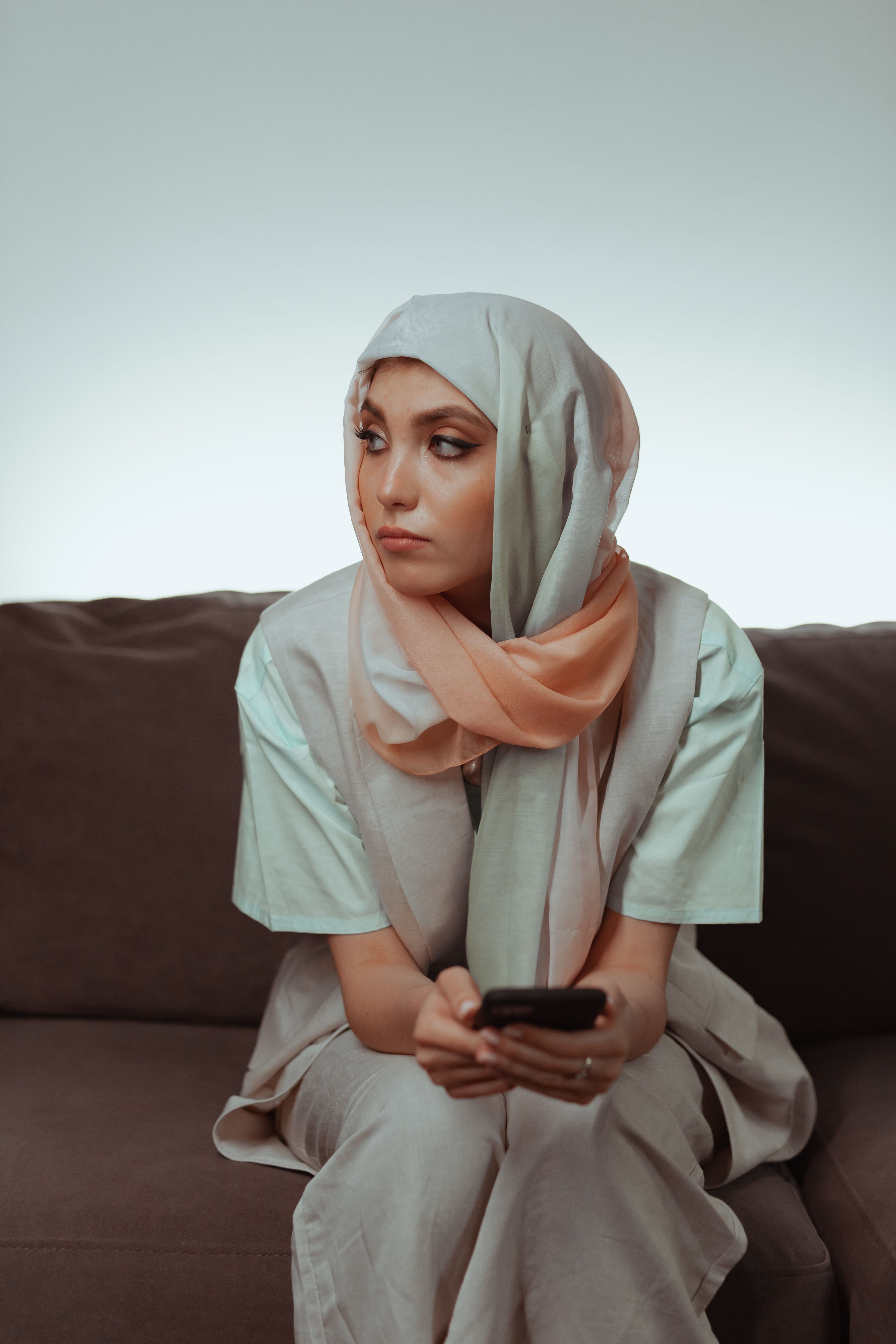 Woman in Gray Hijab Sitting on a Brown Couch and Holding Her Smartphone ...