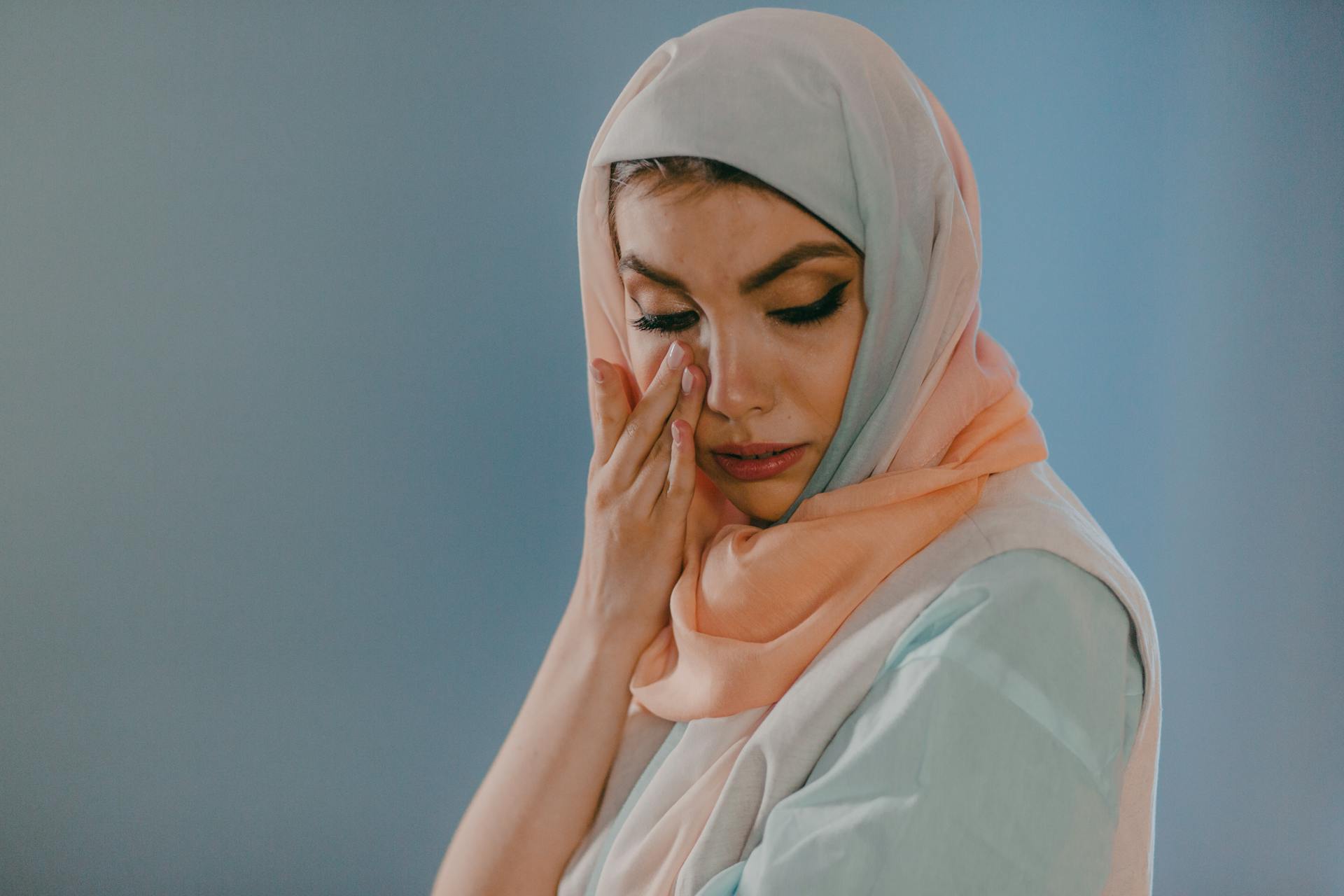 A woman in a hijab wipes tears from her eyes against a blue background.