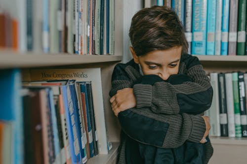 Boy Looking Pensive Wearing a Sweater and Standing Against Bookshelves