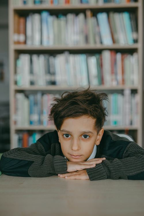 Free A child leaning on desk in a library surrounded by bookshelves, looking thoughtful. Stock Photo