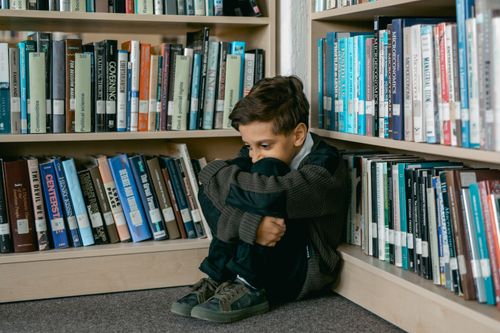 Free Young boy in library appears sad, sitting alone among bookshelves, suggesting themes of solitude and bullying. Stock Photo