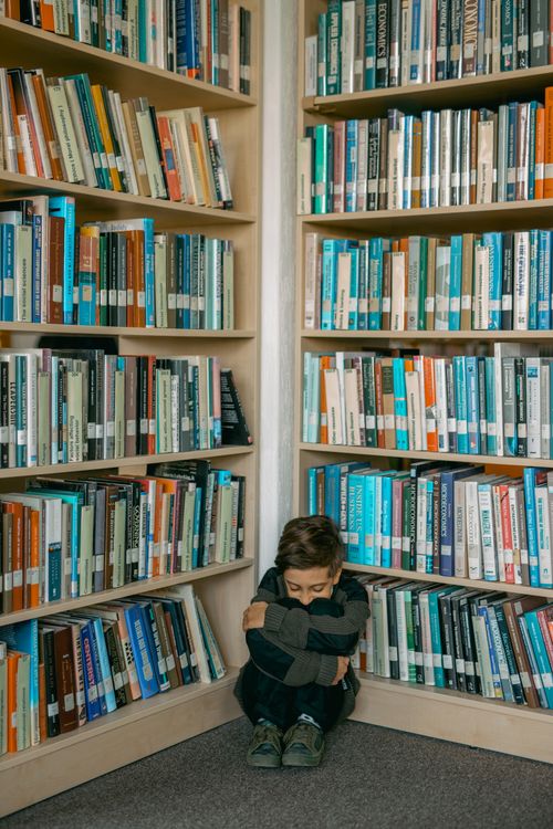 Free A boy sits alone in the corner of a library, surrounded by bookshelves, looking vulnerable and sad. Stock Photo