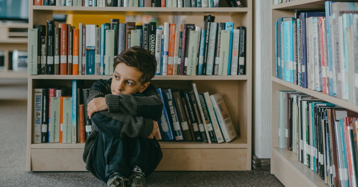 A young boy sits in a library corner, surrounded by bookshelves, deep in thought.