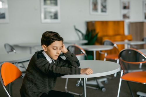 Free Young boy sitting alone at a table indoors, looking contemplative. Stock Photo