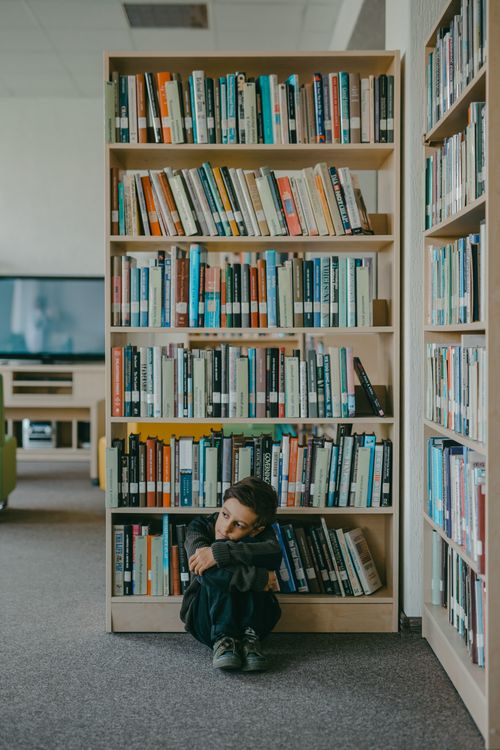 Free A young boy sits alone in a modern library, appearing pensive and introspective. Stock Photo