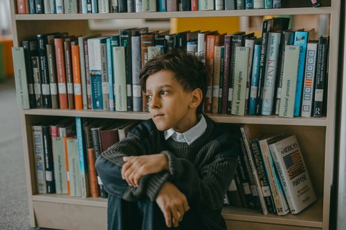 Free A thoughtful young boy sits alone in a library, surrounded by books. Stock Photo