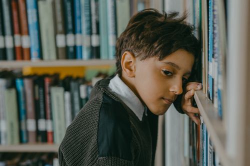 Free A thoughtful boy leans on bookshelves, surrounded by educational books, in a library setting. Stock Photo