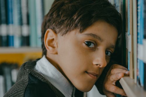 Free Child leaning against a colorful bookshelf, gazing pensively. Stock Photo