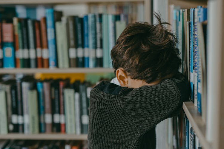 A Boy With Head Down Leaning On A Bookcase