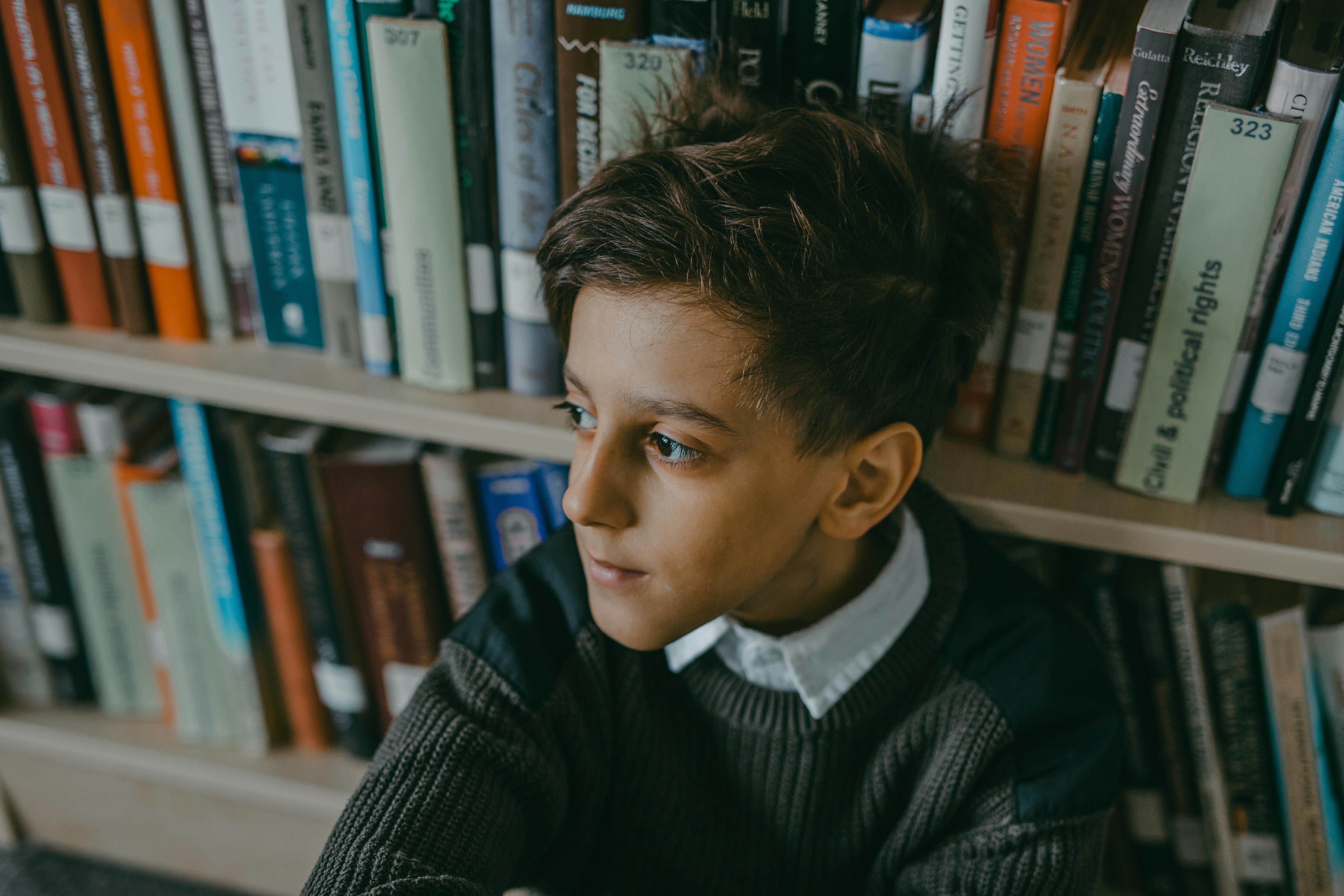 Free A boy seated by a bookshelf in a library looks contemplative and thoughtful. Stock Photo