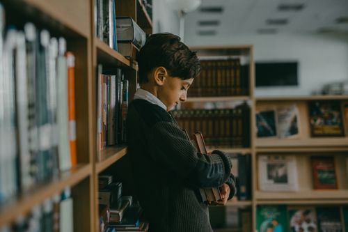 Free A thoughtful young boy leans against a bookshelf, holding books in a quiet library. Stock Photo