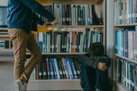 Man in Black Jacket and Brown Pants Standing Beside Books in Shelf
