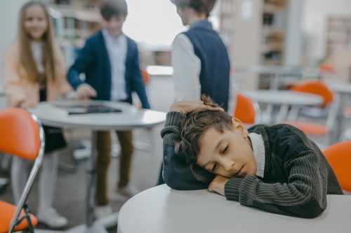 Free A sad child rests on a classroom desk while peers talk, symbolizing loneliness and school exclusion. Stock Photo