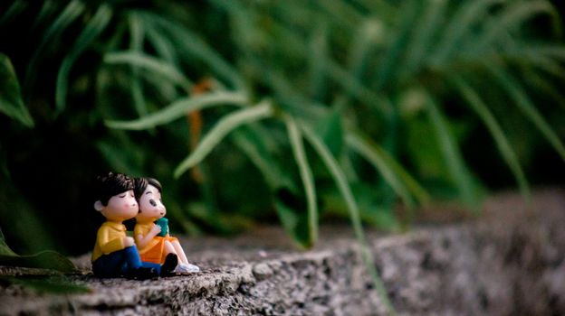 Miniature couple figurines sit on a concrete surface surrounded by lush green plants, creating a whimsical scene.