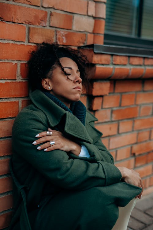 Free Woman in Green Trench Coat Sitting and Leaning Against a Concrete Red Brick Wall Stock Photo