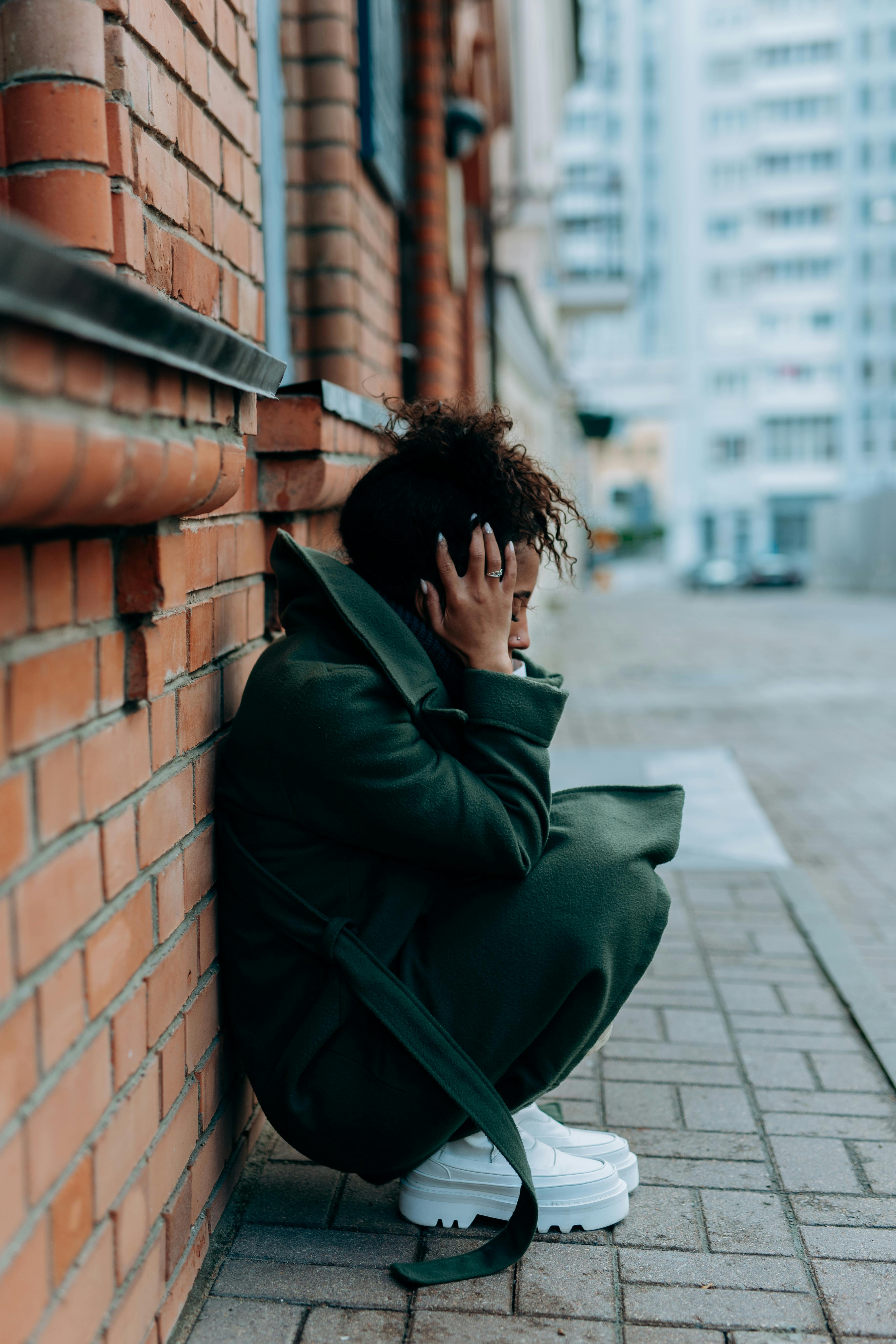 woman in green trench coat sitting and leaning against a concrete red brick wall