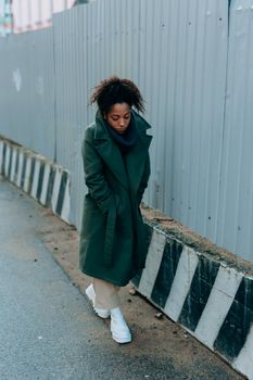 A woman in a green trench coat walks thoughtfully along an urban sidewalk during winter.
