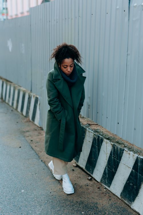 Free A woman in a green trench coat walks thoughtfully along an urban sidewalk during winter. Stock Photo