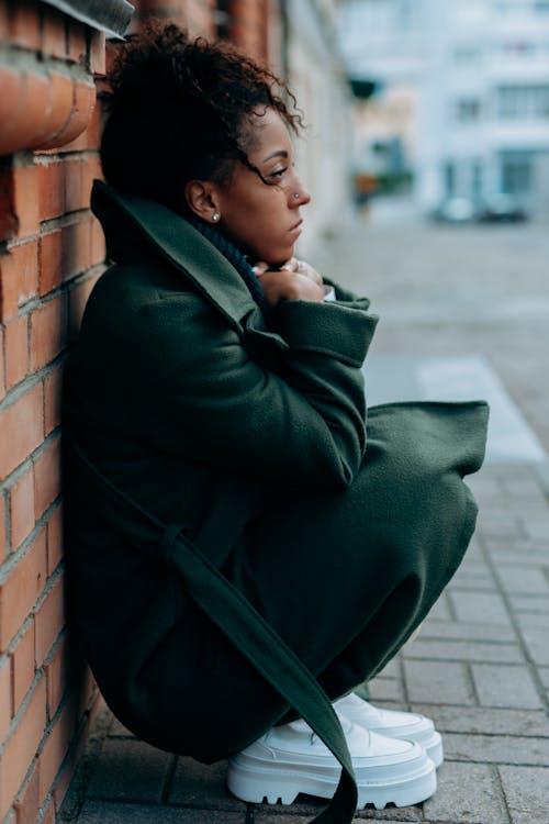 Woman in Green Trench Coat Sitting and Leaning Against a Concrete Red Brick Wall