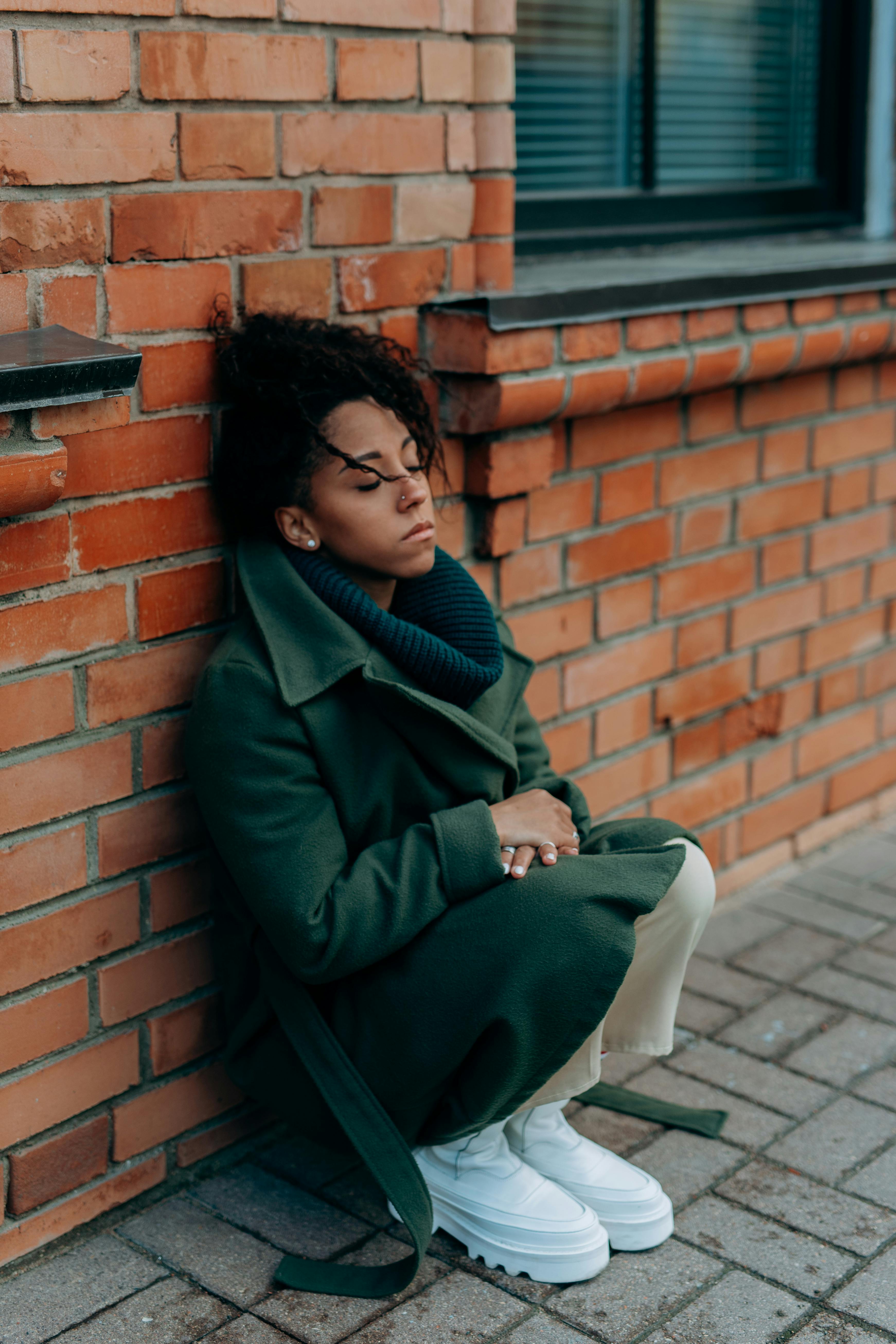 woman in green trench coat sitting and leaning against a concrete red brick wall