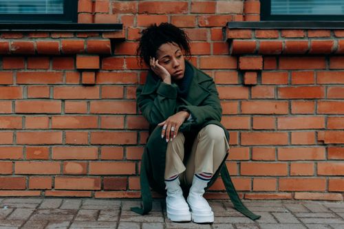 Free A contemplative woman in a green coat sits against a brick wall. Stock Photo