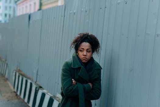 A woman in a green coat walks along an industrial fence on a cold day, reflecting urban fashion.