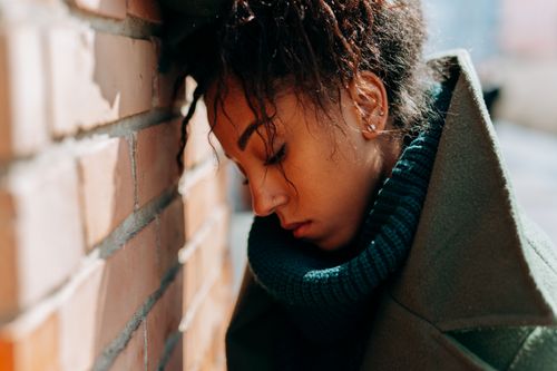 Free Young woman in a green coat leans against a brick wall, eyes closed, capturing introspection and solitude. Stock Photo