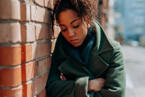Free Young woman in green coat leaning on brick wall, looking thoughtful. Stock Photo