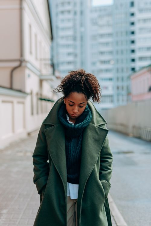 Free A woman in a green coat stands on a city sidewalk, lost in thought, with hands in pockets. Stock Photo