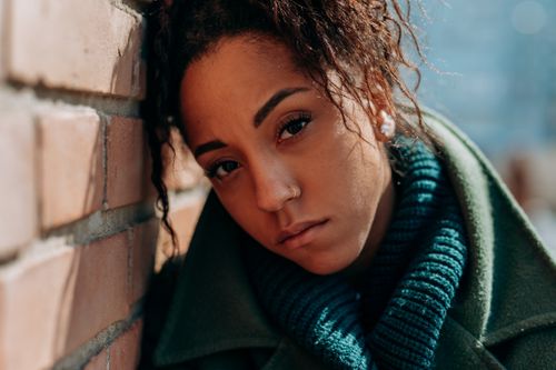 Free A thoughtful woman in a green coat leans against a brick wall on a sunny day. Stock Photo