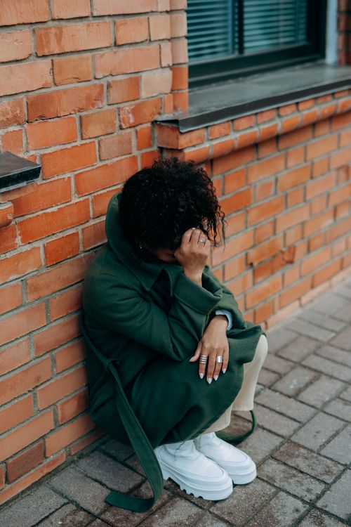 Free A woman in a green coat sits against a brick wall, deep in thought. Stock Photo