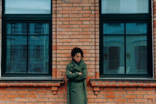 Free A woman in a green coat leans against a brick wall, appearing thoughtful and introspective. Stock Photo