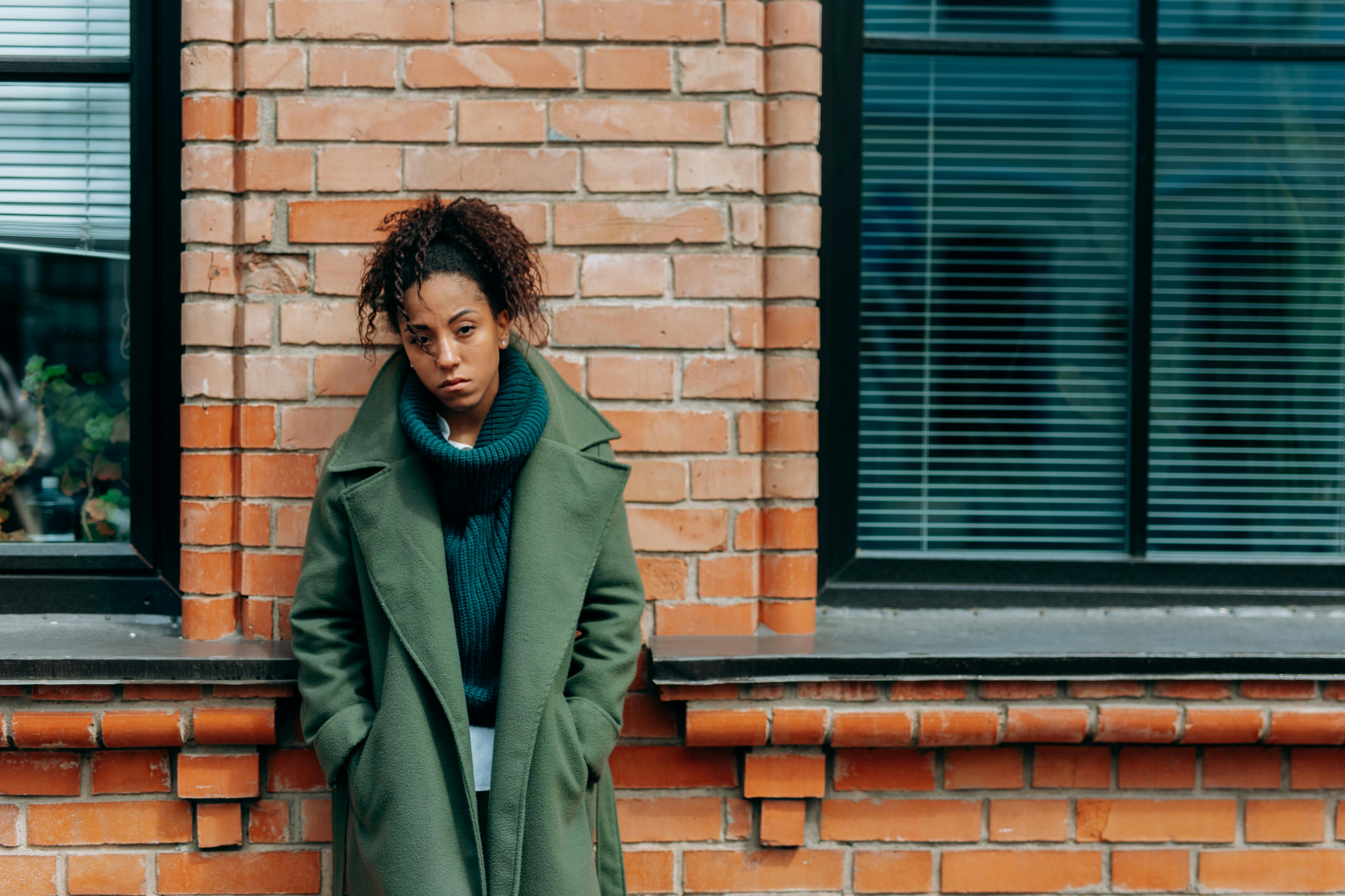 A thoughtful woman in a green coat leans against a brick wall outside during the day.