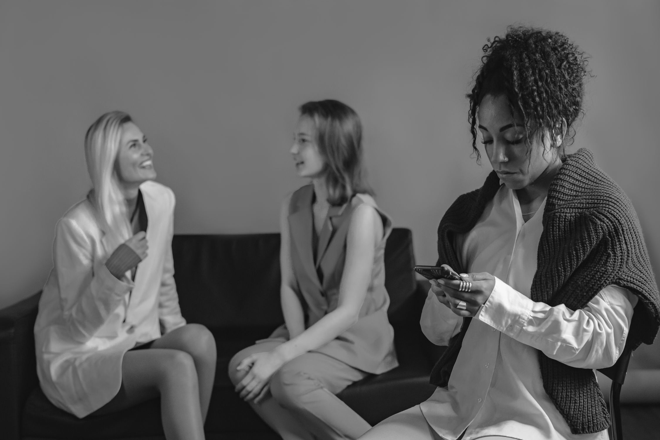 Mulher negra usando celular enquanto sentada afastada de duas mulheres conversando no sofá.