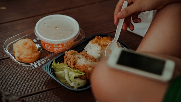 A hand holding a spoon near a meal of fried chicken, rice, and soup.