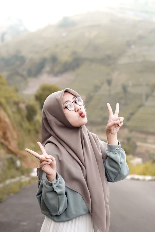 Free A woman in hijab poses outdoors with a peace sign, smiling against a scenic background. Stock Photo