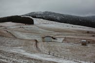 一片白雪皑皑的宁静乡村，田野与谷仓坐落于多云天空下，邻近森林覆盖的山坡