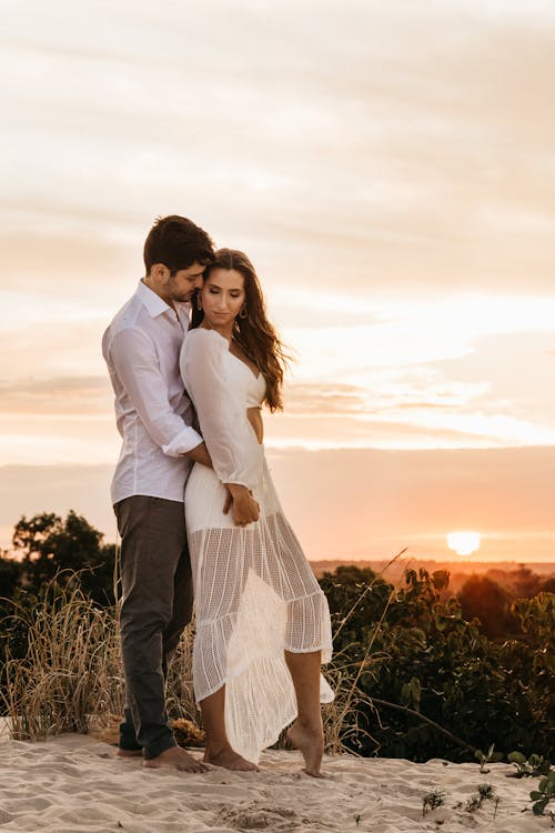 Barefoot lovers standing together on sand against picturesque landscape with sunset and hugging gently