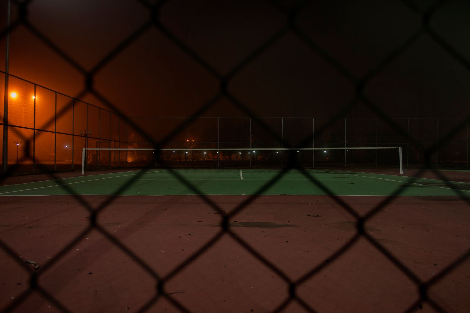 A tennis court seen through a wire fence at night with atmospheric lights