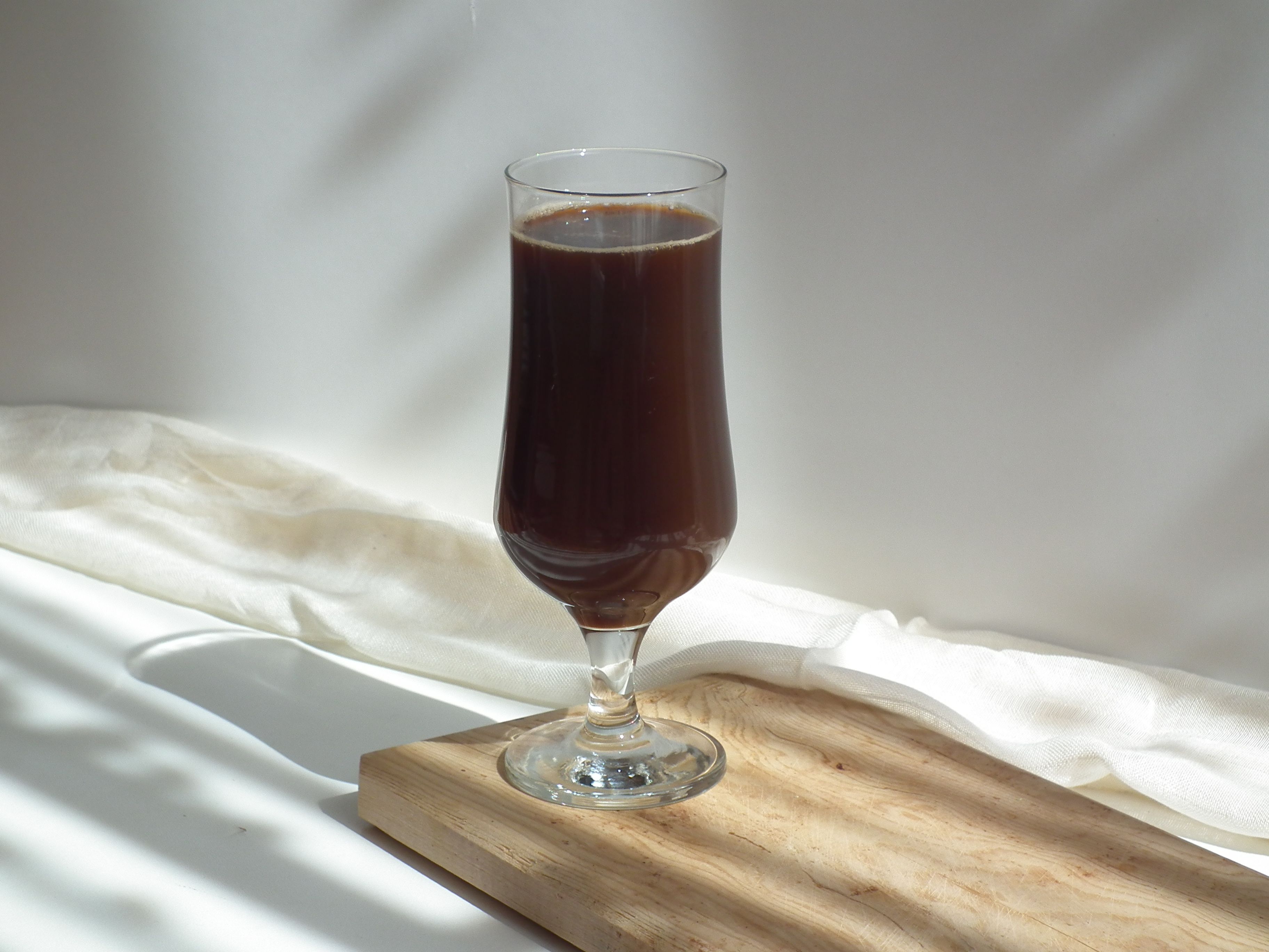 A tall glass filled with a dark beverage placed on a wooden tray against a soft cloth backdrop.