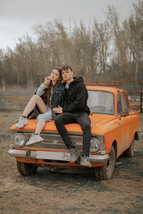 Cool young man embracing girlfriend touching face while looking at camera on vintage automobile parked against autumn trees
