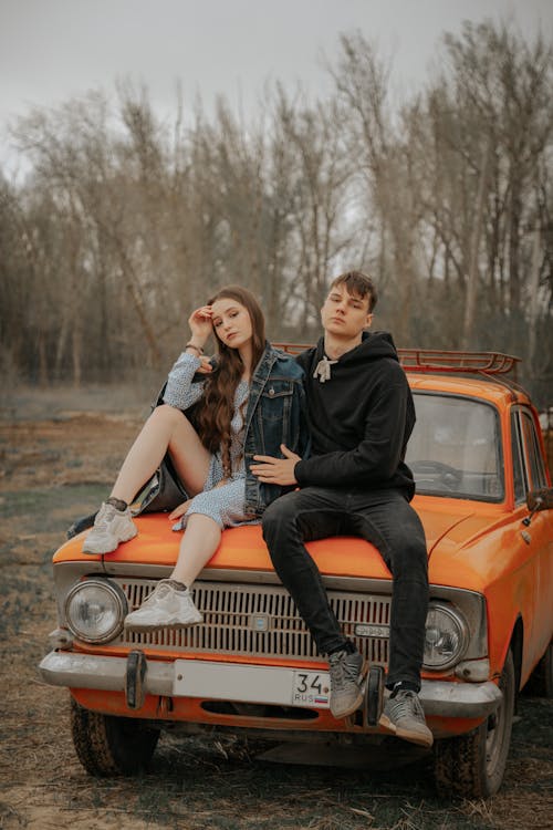 Free Cool young man embracing female partner touching forehead while looking at camera on aged automobile against autumn trees Stock Photo